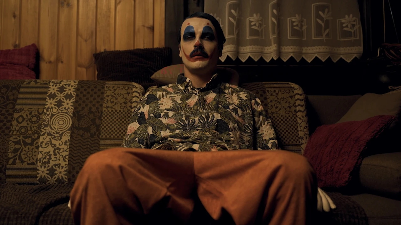 The made-up clown sits legs apart on an old sofa in a Hawaiian shirt and orange trousers.