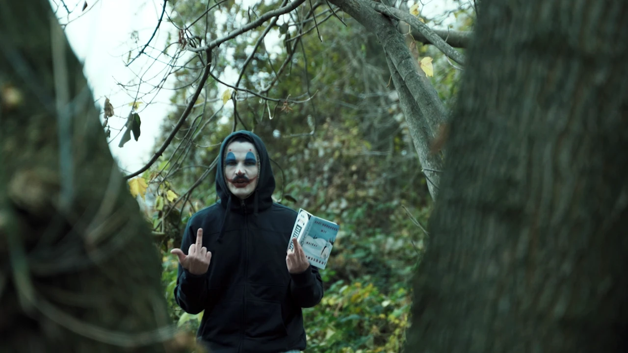 A clown in a hoodie gives the finger in the woods, holding a package in his other hand.