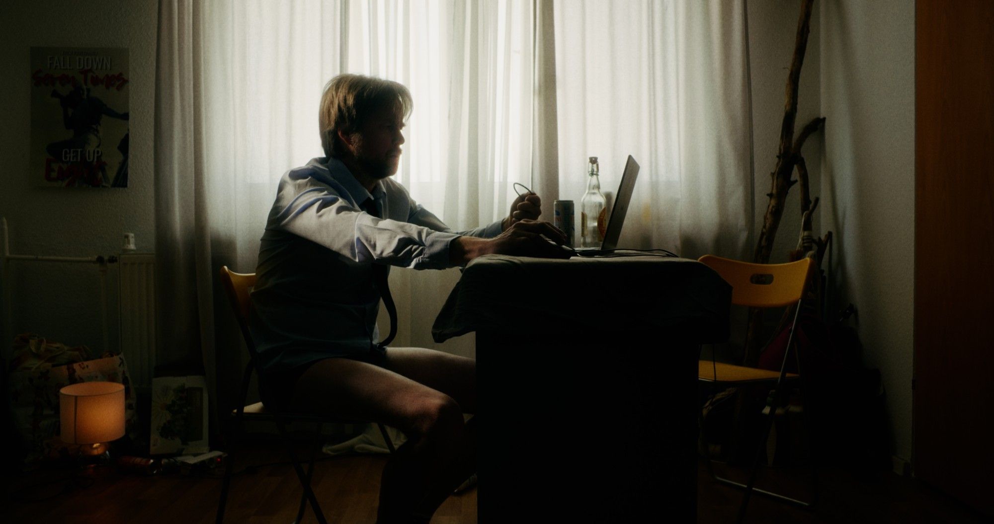 Ein Mann in Hemd und Unterhose sitzt im Gegenlicht vor dem Laptop am Fenster.