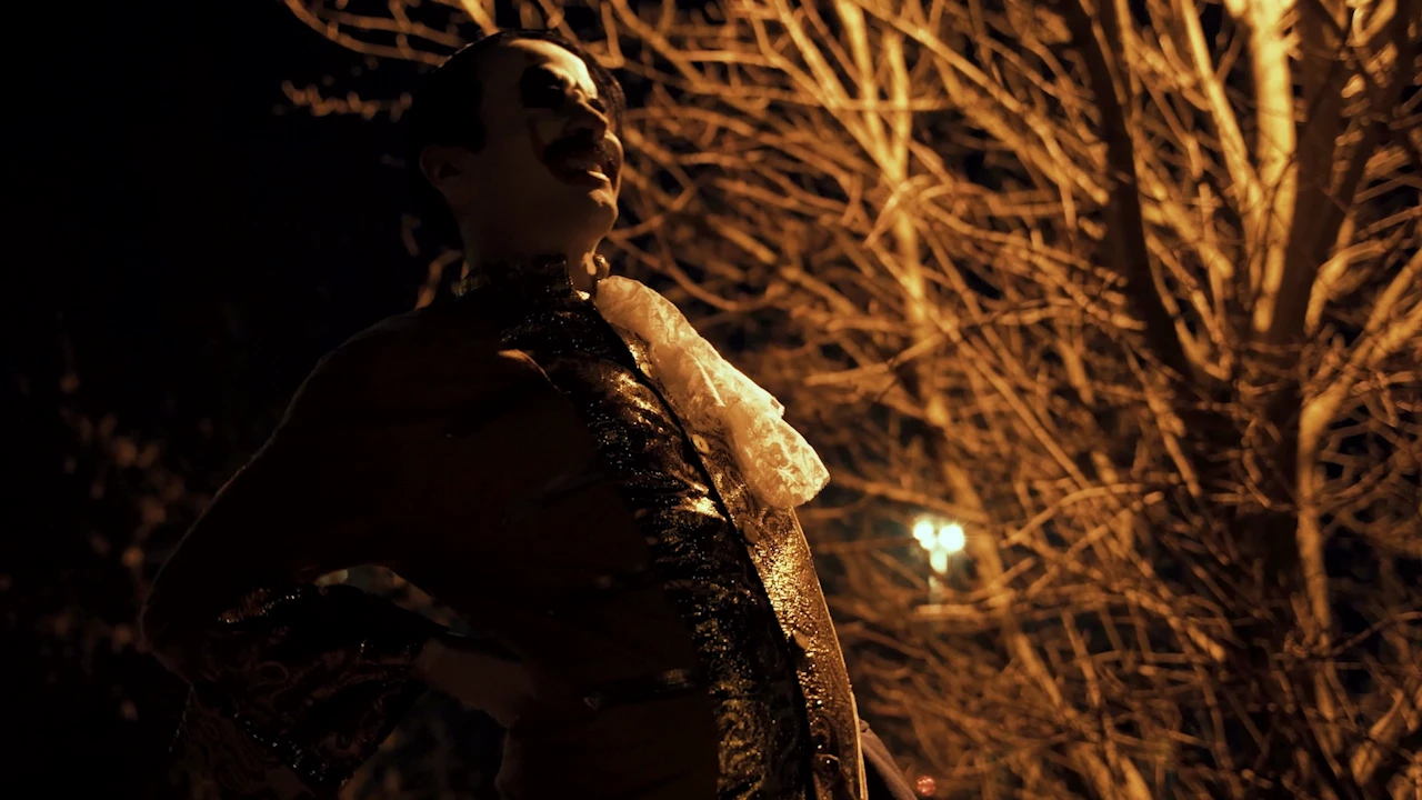 The clown in a shiny costume laughs at night under bare branches lit in yellow.