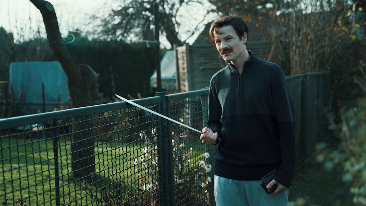 A man with a moustache and a machete stands at a garden fence.