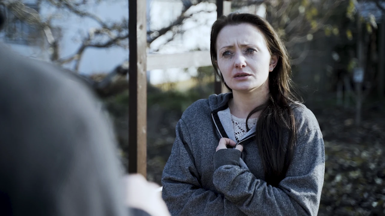 A woman in a grey hoodie speaks with tears in her eyes.