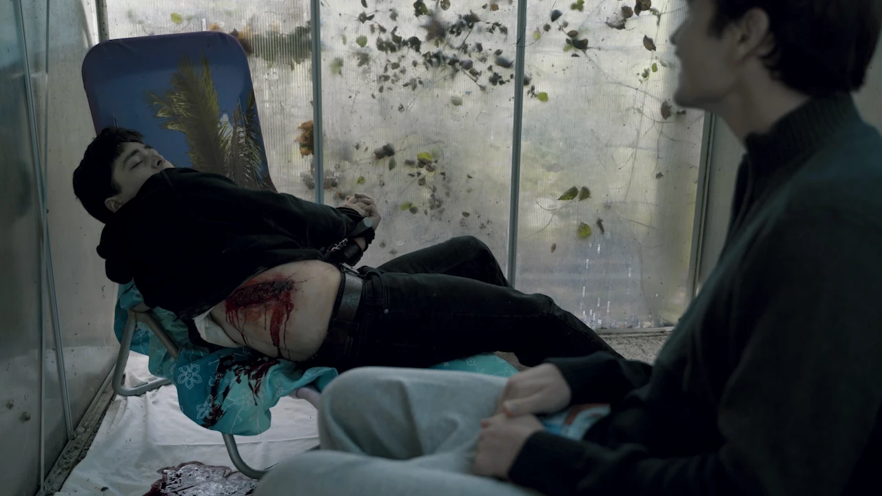 An injured man lies bleeding on a garden chair in a greenhouse, another man sitting beside him.
