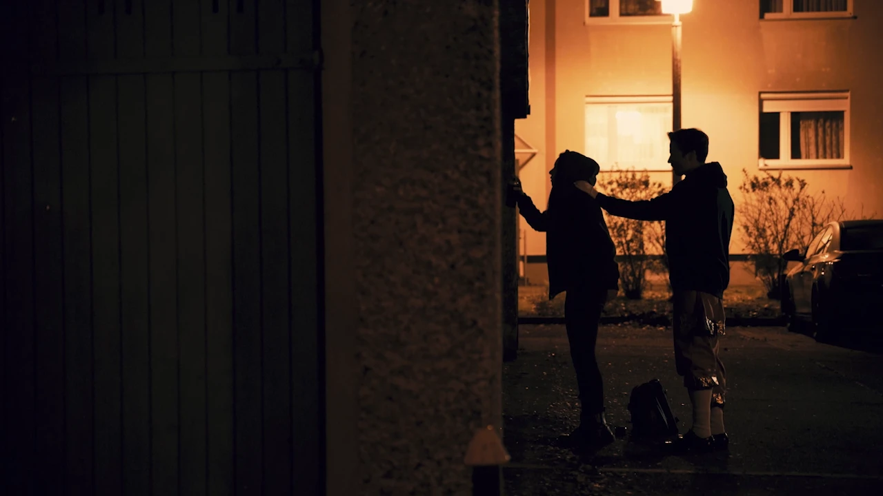 Two silhouettes at night in front of an apartment block, lit by a street lamp.
