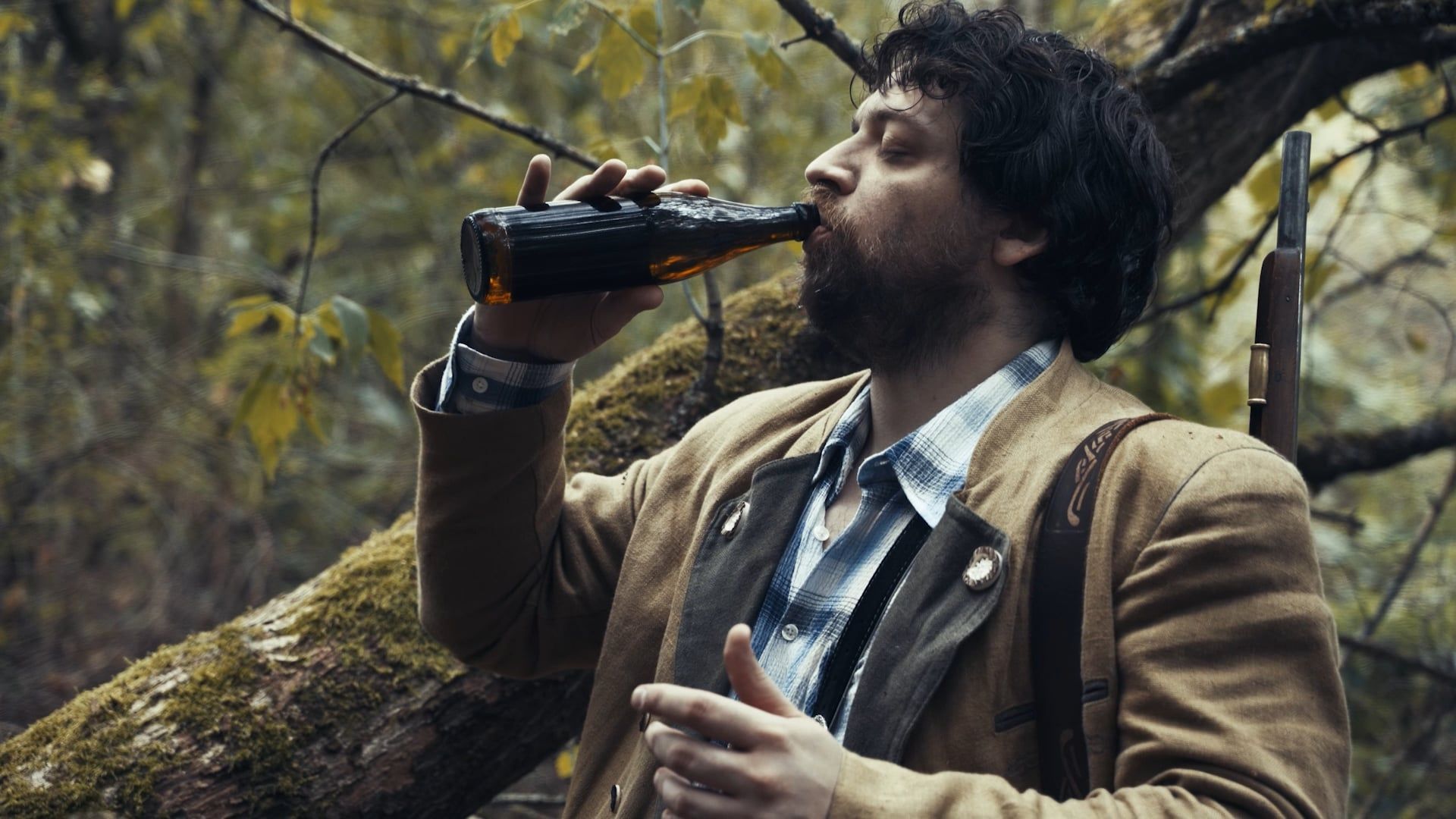 Ein bärtiger Mann in Trachtenjacke, ein Gewehr über der Schulter, trinkt im Wald aus einer Bierflasche.