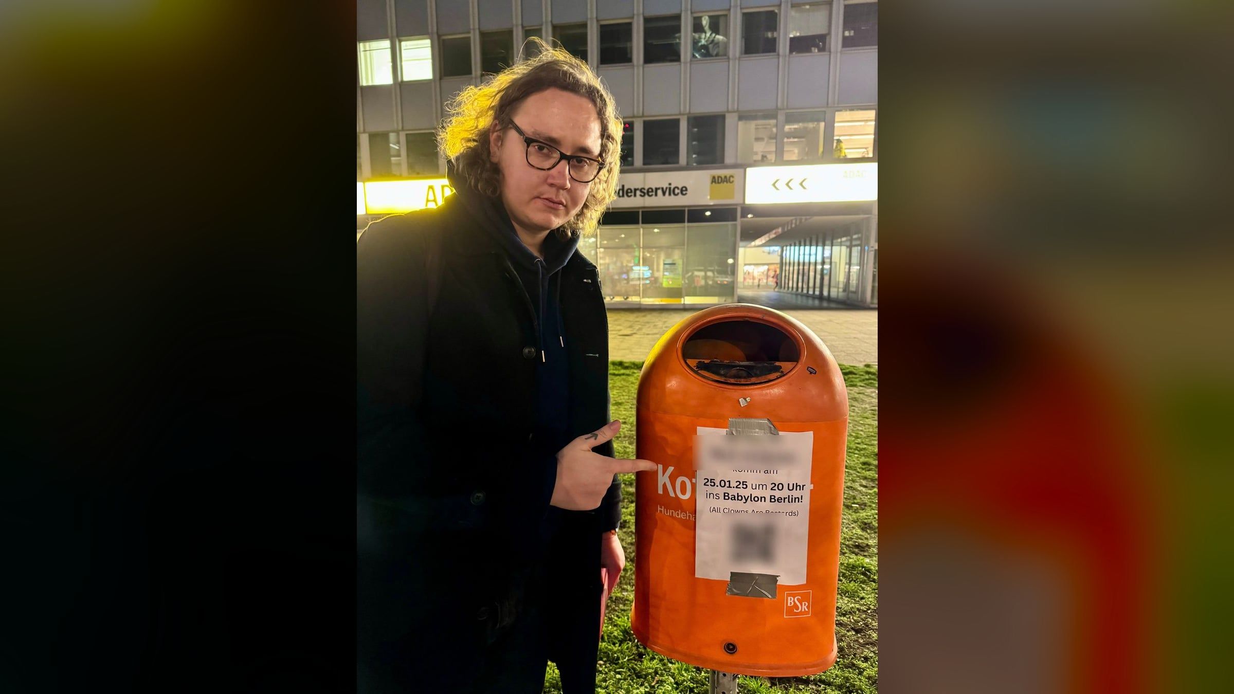 Ein Mann mit Brille zeigt nachts auf einen Zettel an einem orangefarbenen Mülleimer: eine Einladung zu All Clowns Are Bastards im Babylon Berlin.